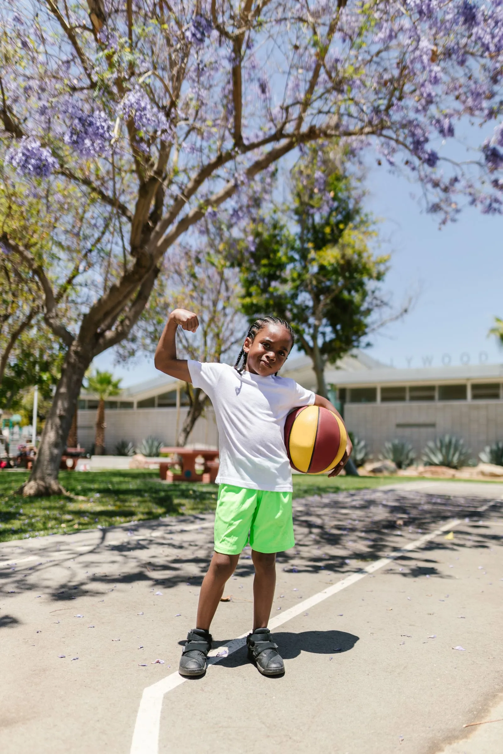 A young player flexing with a basketball under his arm at a sunny park court